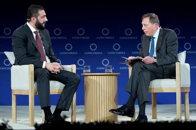 Ahmed Şara, eski CIA Başkanı David Petraeus'a bakarak konuşuyor. Tipik bir sempozyum sahnesinde karşılıklı oturmuş durumdalar.