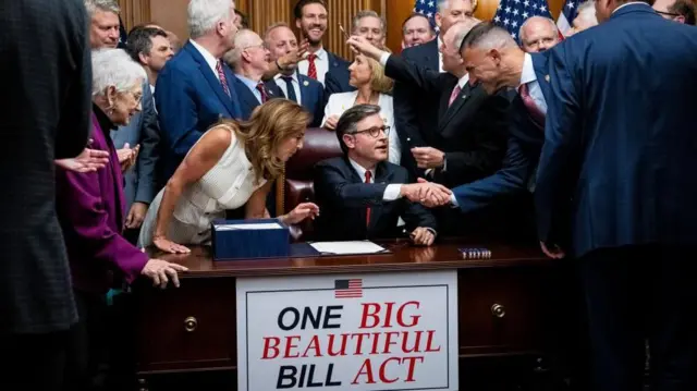 ジョンソン下院議長が机に向かい、周りの共和党議員と握手している。机には、箱に入った法案と、法案を大統領へ送付するための書簡が議長の前に置かれている。議長に、別の議員がペンを渡そうとしている