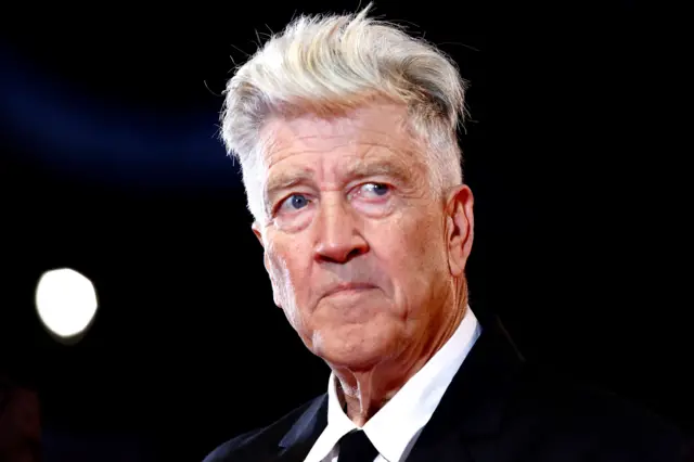David Lynch has light-coloured, voluminous hair styled upward and is wearing a formal black suit jacket with a crisp white dress shirt and a black tie. The background is dark, with a bright circular light source visible on the left side, creating a dramatic contrast and highlighting the hair and upper part of the outfit.
