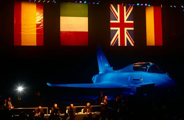 Karanlık bir salonda sahnede savaş jeti var. Arkada ingiltere, İspanya, İtalya, Almanya bayrakları asılı