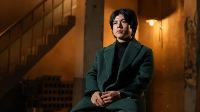 هانيول يجلس على كرسي في غرفة ذات إضاءة خافتة ويرتدي معطفًا أخضر داكنًا بينما يتحدث إلى هيئة الإذاعة البريطانية (بي بي سي).