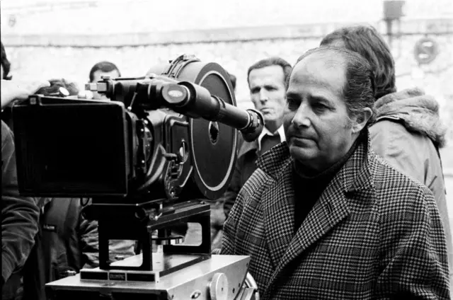 En una foto en blanco y negro, Gillo Pontecorvo está vestido con un abrigo y parado detrás de una cámara de cine de 35mm. Lo rodean otros miembros del equipo de filmación