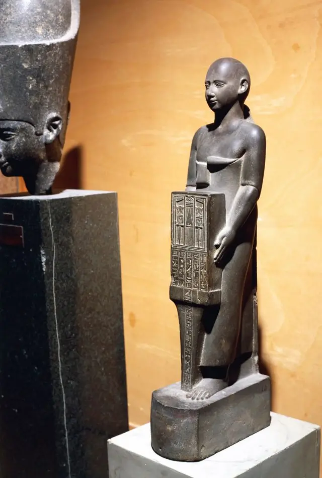 تمثال لأحد الكهنة، من الأسرة السابعة والعشرين، يرتدي رداءً فارسياً ويحمل صندوقاً صغيراً يُظهر واجهة معبد الإلهة نيت في مدينة سايس