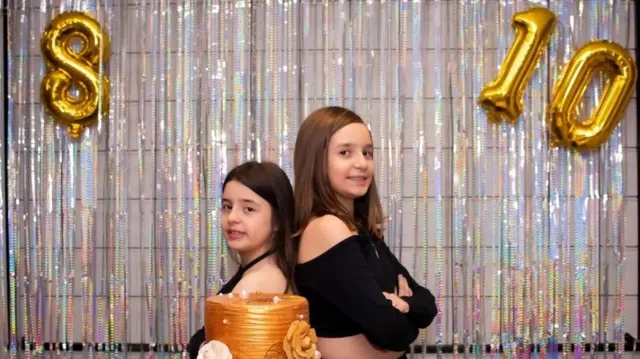 Beatriz ve Lara Bernardi, altın rengi bir doğum günü pastasının arkasında sırt sırta duruyorlar. Arka planda gümüş renkli parti süsleri ve iki altın balon var; birinde 8, diğerinde 10 yazıyor.