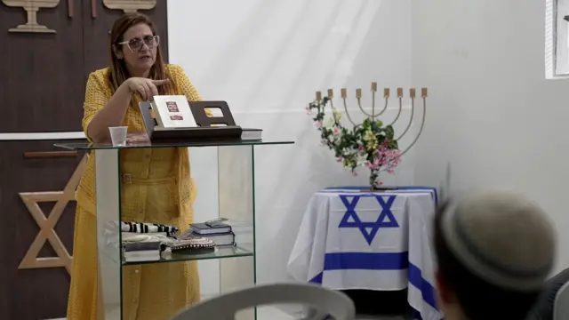 Mulher com roupas amarelas em púlpito; ao lado, há um candelabro e uma mesa com uma bandeira de Israel