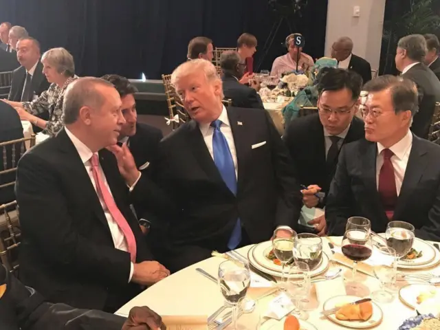 Erdoğan yuvarlak masada Trump'ın yanında oturuyor, masada yemek ve içecekler var birkaç kişi daha görünüyor. İki lider sohbet ediyor.