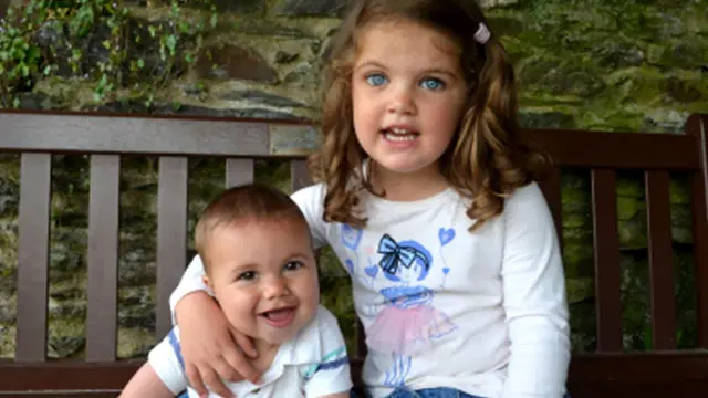 James i Kate siedzą na drewnianej ławce na zewnątrz. Kate obejmuje Jamesa, za nimi znajduje się kamienny mur i zieleń.