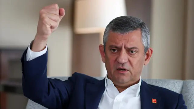 CHP lideri özgür özel'in bir röportajda çekilmiş fotoğrafı. Özel oturuyor, sağ yumruğu havada, beyaz bir gömlek ve lacivert bir ceket giymiş, yakasında chp rozeti var.