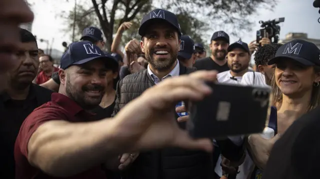 A foto mostra Marçal, um homemjogo do foguete na blazebarba cerrada, sorrindo, rodeado por pessoas, uma delas fazendo uma selfie com ele. Todos estão com um boné azul com a letra M. 