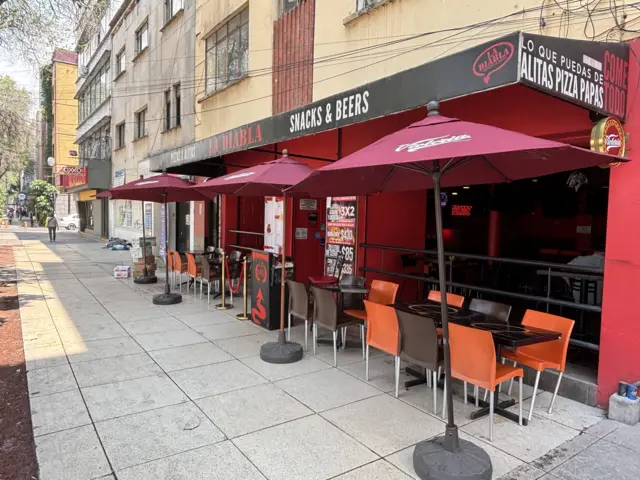 Terraza de restaurante en la acera con mesas y sillas bajo sombrillas rojas, frente a una fachada urbana con letreros.