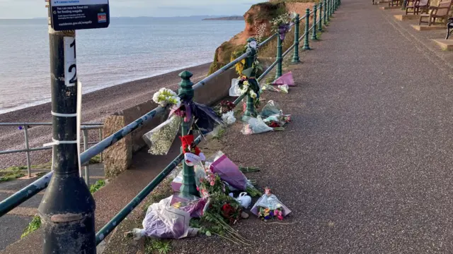 La foto muestra ofrendas florales colocadas a lo largo de las barandillas de la playa de Budleigh Salterton. La playa de guijarros y el mar se ven al fondo, con acantilados a lo lejos.