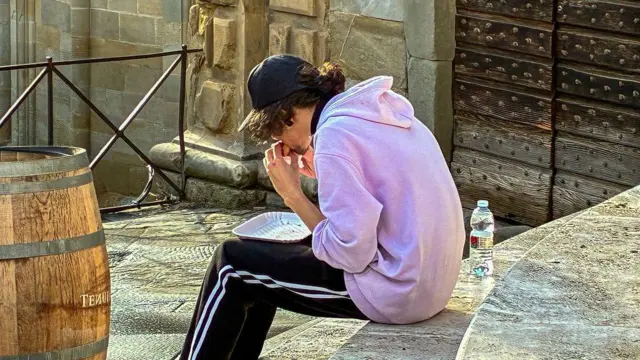 شاب يرتدي سترة وردية اللون وقبعة سوداء يجلس وحيداً على درجات حجرية متآكلة لمبنى تاريخي