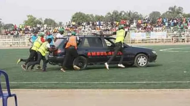 Cinq hommes portant des gilets pare-balles poussent un break noir portant le mot « ambulance » en rouge sur le côté sur un terrain de football.