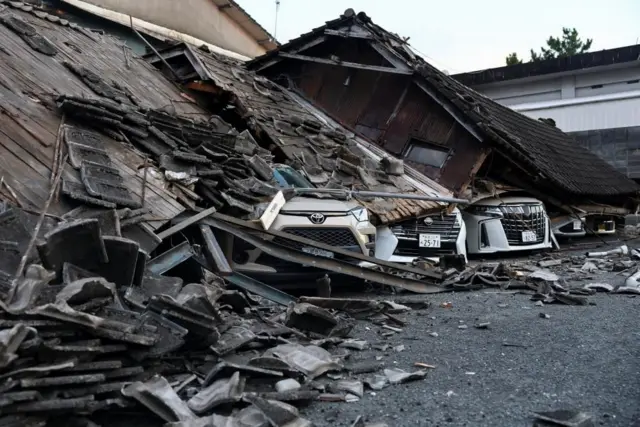 かわら屋根の木造の建物が崩壊し、その下の複数の白い車両にかわらや材木がおおいかぶさっている