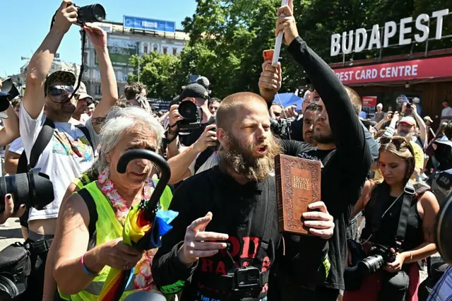 Elinde İncil tutan bir kişi, yürüyüşün Hristiyan inancına aykırı olduğunu iddia ediyor. Sarı sakalları uzamış.