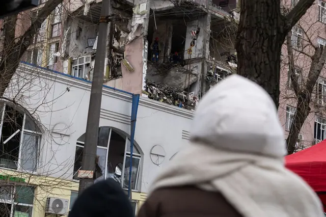 Зруйновані два поверхи багатоповерхового будинку: стіни зруйновані, оголені перекриття, всередині хаос із уламків та речей. На балконі працюють рятувальники у захисному одязі. На передньому плані – людина в білому капюшоні та шарфі, яка дивиться на руйнування.