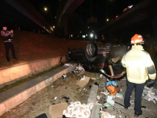 야간의 고가도로 아래 사고 현장으로, 검은색 승용차 한 대가 완전히 뒤집힌 채 바닥에 놓여 있다. 차량 주변에는 부서진 부품과 각종 물건들이 흩어져 있다. 형광색 안전복을 입은 구조대원이 차량을 살펴보고 있다.