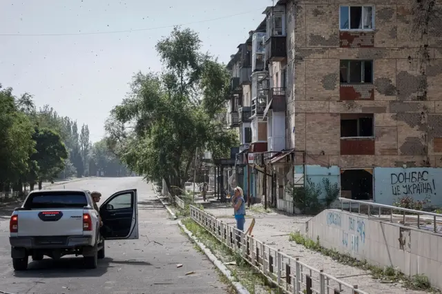Žena stoji na ulici ispred stambenog bloka, razgovara sa policajcem koji se nalazi u zasutavljenim kolima na ulici, region Donjeck, jul 2024. 