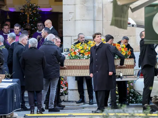 Люди вносят в церковь гроб, сделанный в стиле плетеной корзины. Он украшен желтыми и оранжевыми цветами.