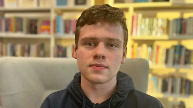 Liam has short, brown, wavy hair and is wearing a grey hoody. He is sitting in a grey high-backed armchair in a library and looking directly into the camera. 