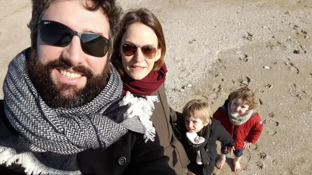 Kumlu bir plajda çekilmiş yakın plan bir özçekimde dört kişi görülüyor. Ön plandaki iki yetişkin güneş gözlüğü, mont ve atkı takıyor. Aşağıda duran iki küçük çocuk kameraya bakıyor; biri kırmızı, diğeri siyah mont giyiyor. Arka plandaki kumlar deniz kabukları ve ayak izleriyle kaplı