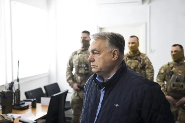 У світлому приміщенні стоїть кілька людей у камуфляжній формі та спорядженні, розташованих позаду центральної фігури Орбана, вдягненого у темну стьобану куртку. У кімнаті видно стіл із раціями, паперами та офісними стільцями, а крізь вікно надходить яскраве природне світло. 