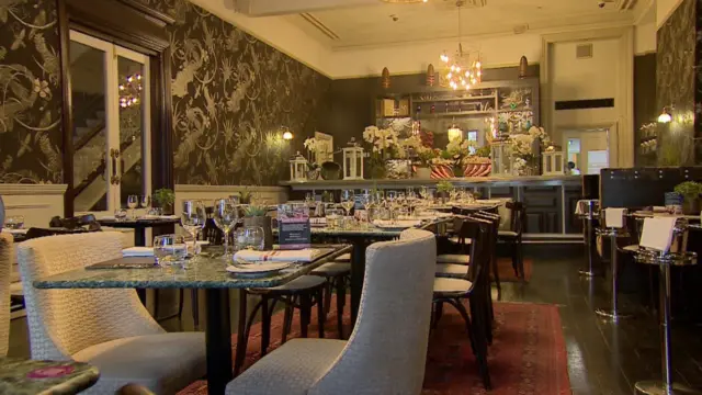 Comedor de un restaurante. Las paredes están decoradas con papel pintado oscuro con estampados, mientras que el techo es blanco. Una lámpara cuelga del techo sobre la barra, al fondo del local. En primer plano, se encuentran mesas de mármol verde con copas de vino y platos. Hay sillas de tela beige/clara junto a las mesas. El suelo de madera oscura está cubierto por alfombras rojo oscuro.