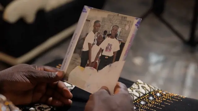 Close shot of Mrs Agbro hands wey hold foto of her chidldren. Di late Favour and her sister Fith wit dia brother pose wit wetin resemble birthday cake.