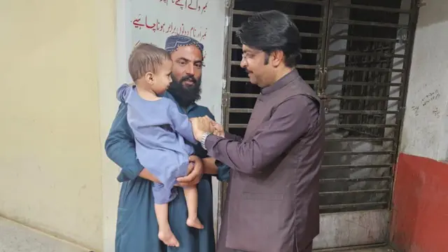 ဦးထုပ်ဆောင်းထားတဲ့ ဂျလီအယ် အာမက်ဒ်ဟာ ပိုလီယိုရောဂါခံစားနေရတဲ့ သားဖြစ်သူ ဂျဝက် ကို ချီပိုးထားရင် နောက်ထပ်အမျိုးသားတဦးနဲ့စကားပြောနေစဉ်