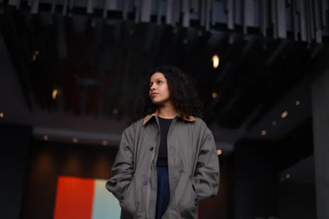 Arianna de Souza-García vistiendo una chaqueta holgada de color gris; está de pie bajo un techo moderno con paneles oscuros y luces tenues, frente a un fondo interior con un rectángulo iluminado.