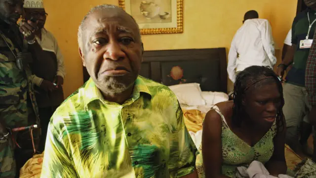 Laurent Gbagbo et Simone Gbagbo sont assis sur le bord d'un lit, l'air abattu. Derrière eux se tiennent plusieurs personnes, dont un homme en uniforme militaire.
