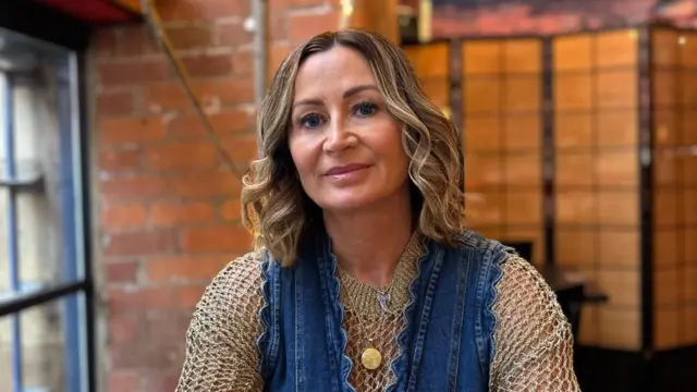Person seated at a restaurant table, arms resting on the surface, wearing a denim dress layered over a textured long-sleeve top, with a watch, rings, bracelets and a round pendant necklace. Sunglasses lie on the table to the left. Behind, a brick wall and decorative branches with pink blossoms and small warm fairy lights. A wooden room divider and a wall mural with a sunset scene appear in the background. Soft indoor lighting throughout.