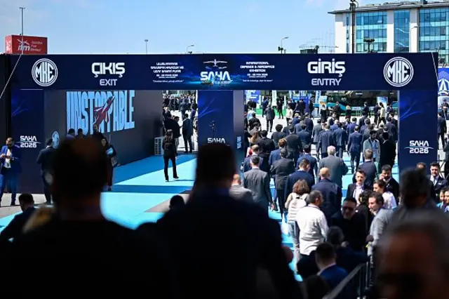 Saha expo'nun giriş ve çıkış yazılı bir geçişi yanyana girişten çok sayıda resmi giyimli insan geçiyor.