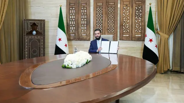 Suriye'nin geçici devlet başkanı Ahmed Şara, geniş bir masada oturmuş, elindeki 18 Ocak anlaşmasını gösteriyor. Arkasında Suriye bayrağı var. 