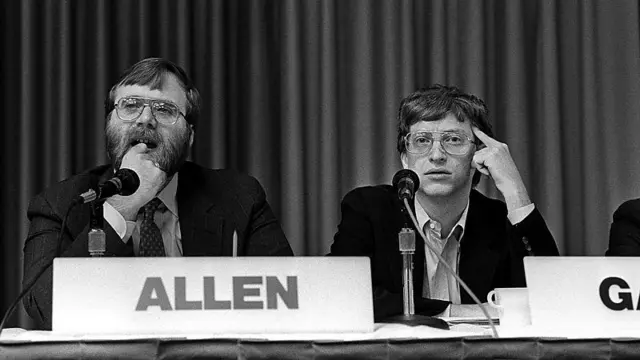 Paul Allen e Bill Gates sentados em uma mesa de evento, ouvindo atentamente