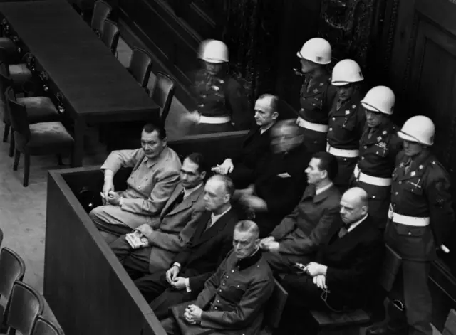 हर्मन गोरिंग (नाझी हवाई दलाचा माजी प्रमुख), रुडॉल्फ हेस (माजी डेप्युटी चान्सलर), जोआकिम वॉन रिबेनट्रॉप (माजी परराष्ट्र मंत्री), आणि विल्हेल्म कीटेल (माजी नाझी लष्करप्रमुख) यांचा फोटो, ते न्युरेंबर्ग पॅलेस ऑफ जस्टिसच्या कोर्टरूममध्ये बसलेले होते