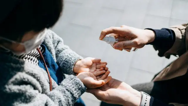 Criança com máscara recebe álcool gel para limpar as mãos