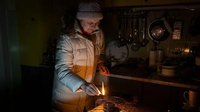 Жінка в зимовому одязі без світла в квартирі намагається розпалити газову пічку