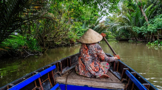 Starsza kobieta w długiej wzorzystej sukience i tradycyjnym stożkowym kapeluszu siedzi w niebiesko-czarnej łodzi i wiosłuje. Łódź płynie w dół rzeki. Oba brzegi są gęsto porośnięte roślinnością. 