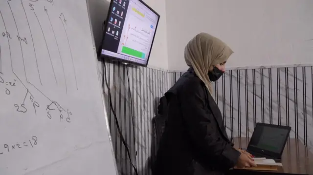 معلم کتابی را بالای میز و در پهلوی یک لپ‌تاب است بازکرده و به آن نگاه می‌کند. پشت سر او یک تلویزیون و تخته سفید است. 