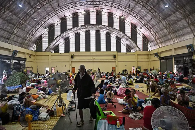 Seorang pria tua berjalan dengan tongkat di antara orang-orang lain di sebuah GOR.