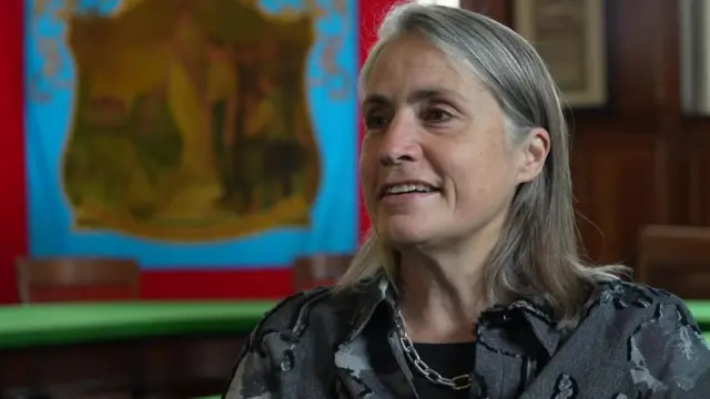Fiona Hill smiling to the left of the camera. She has shoulder length grey hair and is wearing a grey shirt and silver chain necklace. She looks to be sitting in front of a blue and red miner's banner which a painting in the middle.