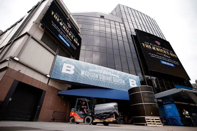 Do lado de fora do Madison Square Garden, uma pessoa dirige uma empilhadeira transportando equipamentos para dentro da arena. Ao fundo, é possível ver a fachada e a identificação do local