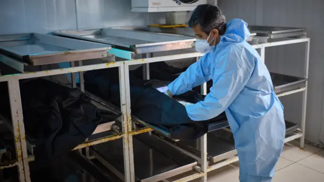 A male hospital employee wearing a protective blue suit and white face mask checks the body of a medical member of staff who died from Covid. This is taking place inside a morgue in Rasoul-e-Akram hospital in western Tehran on 8 December 2020. The body is in a black bag that is placed inside an open rack. Another body is in a similar bag in the compartment next to where the worker is checking the body.