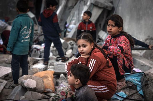 Crianças sentadas em meio aos escombros de edifícios destruídos após bombardeio israelense em Rafah, em 29 de dezembro