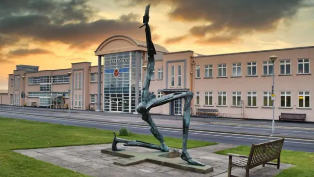 La fachada del aeropuerto de Ronaldsway: un edificio largo y de color claro con una gran entrada acristalada. La estatua de bronce de "El hombre de las tres piernas", obra de Bryan Kneale, se encuentra en el exterior y se puede ver en primer plano.