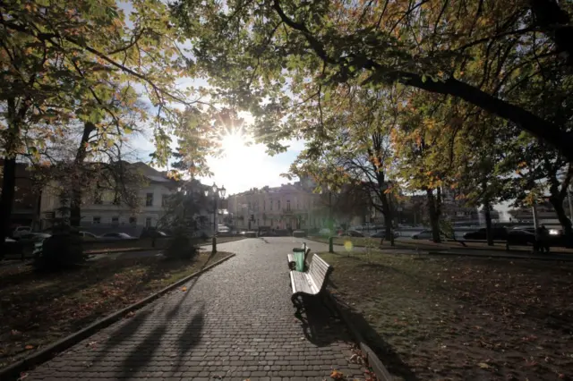 Вимощена бруківкою доріжка, вздовж якої стоять дерев’яні лавки та зелені урни. У тлі – старовинні будівлі, автомобілі припарковані вздовж вулиці. Над доріжкою нависають гілки дерев із зеленим та жовтуватим листям, крізь які пробивається яскраве сонце.
