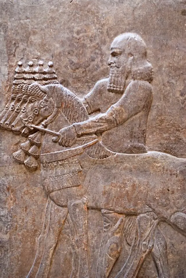 La figura de un hombre esculpida en piedra. De cuerpo entero, se encuentra de perfil con un caballo. A lo largo se ven más caballos.