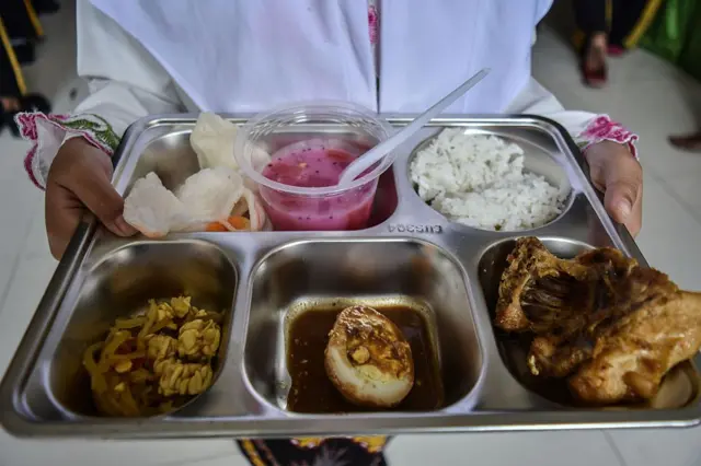 Seorang siswa mengangkat ompreng berisi berbagai jenis makanan berat dari nasi, telur setengah, dan tempe orak, tanpa menunjukkan wajah.