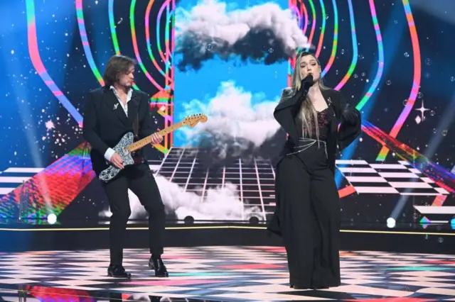 Момак и девојка на сцени, обоје обучени у црно, она пева, он свира гитару